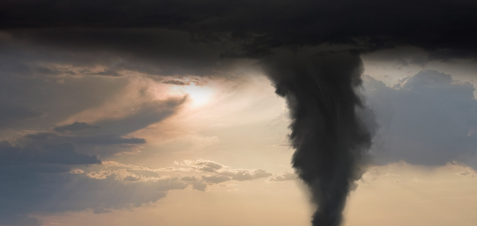 The funnel of a tornado and it's dark clouds overshadowing a  lighter sky and sun in the distance,